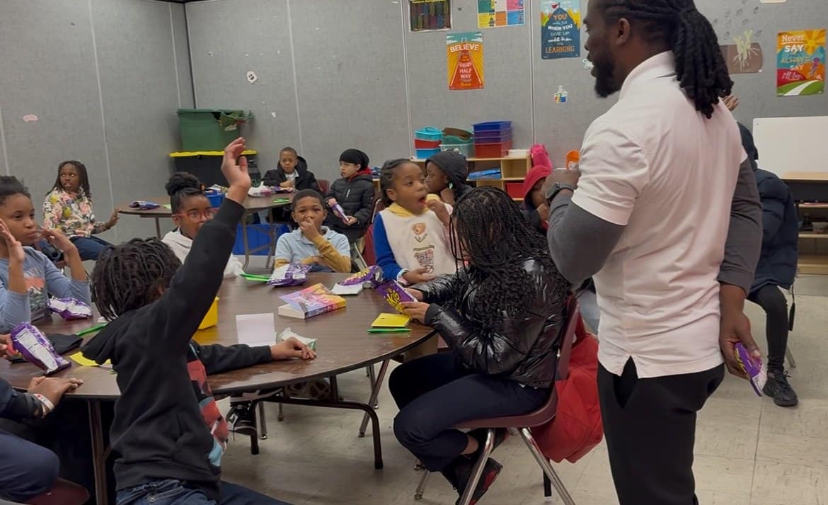 An instructor leads an elementary school activity while students sit around a classroom table.