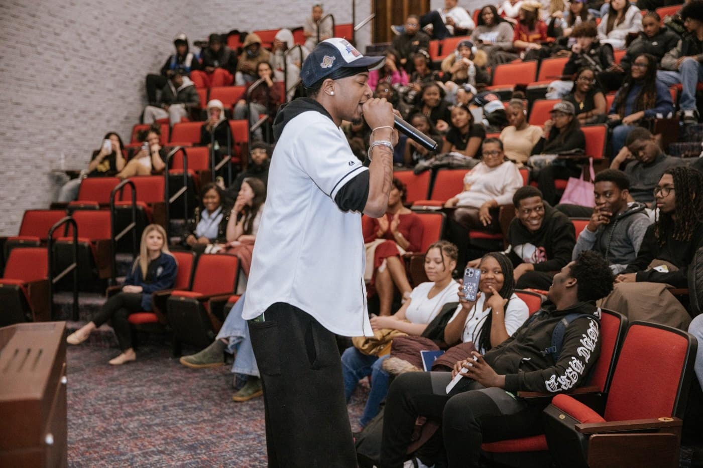 A performer speaks into a microphone while engaging students in the auditorium.